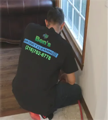 A Ben's Air Duct Cleaning technician in a branded shirt cleaning a vent by a window